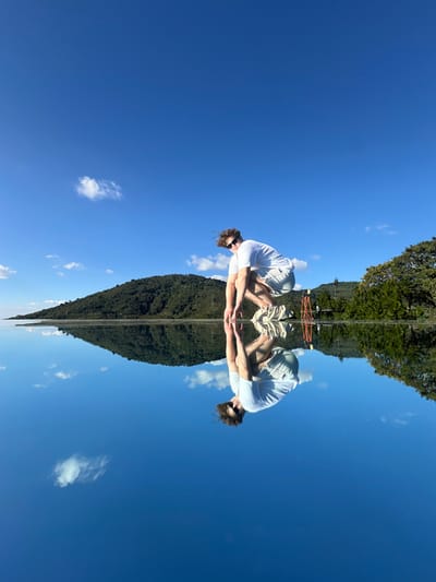 A surreal, symmetrical image shows a mirrored scene. Two figures wearing white shirts and sunglasses sit facing each other, their hands touching. Above and below them, a tall, dense line of trees stretches to the top and bottom edges. A road bisects the tree line. Blue sky with scattered clouds fills the spaces around the central element, enhancing the dreamlike quality of the image.
