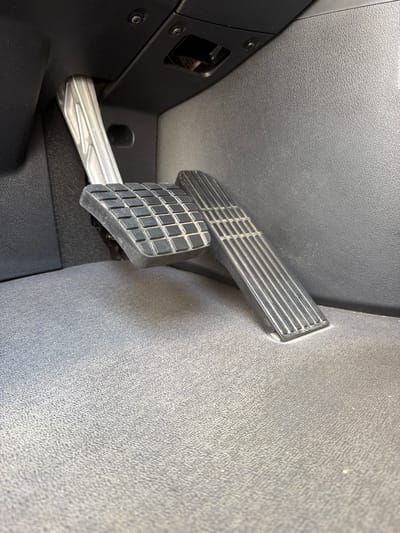 The image shows the interior of a vehicle, specifically the driver's side footwell. Two pedals are visible: a wider, ribbed pedal on the right, and a narrower, grid-patterned pedal on the left. The pedals are made of metal and rubber. The floor of the footwell is covered in dark gray carpet, which appears dusty. The surrounding interior surfaces are made of black and dark gray plastic. A silver, ribbed metal arm extends upwards from the left pedal towards the dashboard. The scene is lit by overhead light, casting soft shadows. There are no people or any indication of activity. This image was taken in Lebanon, United States of America.