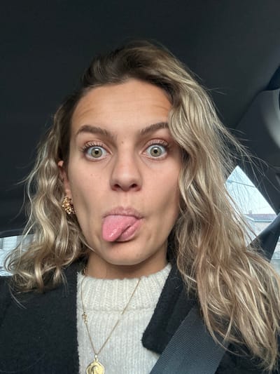 A young woman with blonde wavy hair is making a silly face at the camera. Her eyes are wide, and she has her tongue sticking out. She is wearing a white sweater under a black coat. A gold necklace and earrings are visible. The background suggests she might be in a car.