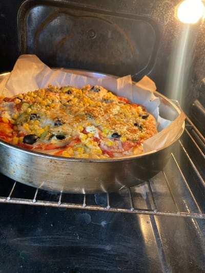 A round pizza, topped with melted mozzarella cheese, yellow corn kernels, sliced black olives, green bell peppers, and white cheese crumbles, rests on a red sauce base. The melted cheese exhibits slight browning in areas. The pizza is contained within a circular metallic baking pan, lined with crumpled parchment paper around its perimeter. The pan is positioned on a metal wire oven rack inside a dark oven interior. An illuminated incandescent oven light is visible in the upper right section of the oven. No individuals are present. The scene is located within a kitchen in Raduil, Bulgaria.