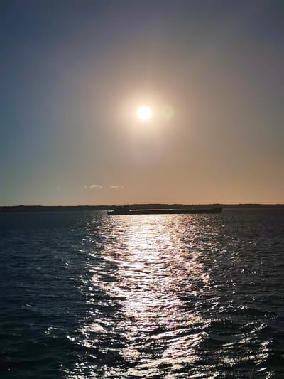 The image depicts a serene seascape at sunset, with the sun casting a shimmering golden path across the water. A cargo ship is visible on the horizon, silhouetted against the warm glow of the setting sun. The sky is a gradient of blues and oranges, with a few wispy clouds. The water is calm, with gentle ripples reflecting the sunlight. The foreground is dominated by the dark blue water, with streaks of light breaking through the surface. The background shows a dark, tree-lined horizon. The location is stated as Voronino, Russia, although the image itself does not provide any specific visual cues to confirm this location.
