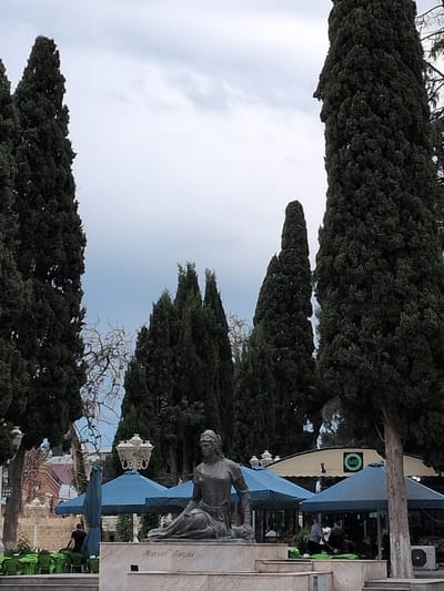 In the city of Ganja, Azerbaijan, a bronze statue of a seated woman is positioned in the foreground, her gaze directed slightly to the viewer's left. The statue rests on a marble pedestal inscribed with the name "Nizami Ganjavi". Behind the statue, several large cypress trees dominate the scene, their dark green foliage creating a dramatic backdrop against a cloudy, overcast sky.

The middle ground features a seating area with bright green chairs arranged around tables that are covered by blue umbrellas. A person in a black shirt is visible in the distance, interacting with the chairs. Further back, a building with a light-colored roof and a circular logo with the letters "MB" can be seen. Several other people are present under the umbrellas. The overall impression is of a public space, possibly a park or a cafe, with the statue serving as a prominent focal point. The presence of the statue of Nizami Ganjavi, a celebrated 12th-century Persian poet who lived and worked in Ganja, strongly suggests the location is Nizami Park.