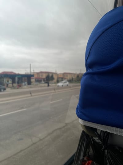 The view is from inside a vehicle, looking out onto a street with buildings on either side. The foreground includes a blue athletic garment with a white trim, partially obscuring the view. A black plastic bag is also visible. The street has multiple lanes of traffic with vehicles visible. The buildings along the street appear to be a mix of residential and commercial properties, possibly a shopping area. The sky is overcast. While the location is purported to be Karmana, Uzbekistan, there is no definitive evidence within the image to confirm this claim.