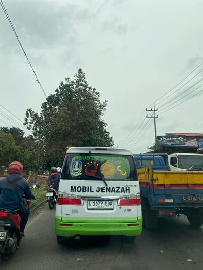 A white and green "Mobil Jenazah" (Funeral Car) with license plate L 1677 BDA is positioned on a road. Stickers on its back include "RSUD Dr. Soetomo" and other logos. A motorcycle is right behind the car. A large truck is visible alongside the car, next to a "Platinum" sign. Overhead, power lines traverse the scene against a cloudy sky.