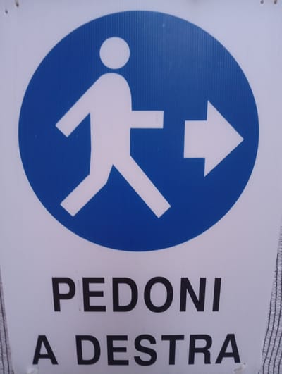 The image is a close-up, eye-level shot of a white sign with a blue circular graphic and black text. The graphic depicts a stylized white figure of a person walking with their left leg forward, and an arrow pointing to the right. Below the graphic, the text reads "PEDONI A DESTRA". This translates from Italian to "Pedestrians to the right". The sign is likely located in a public area in Civitanova Marche, Italy, indicating a designated pedestrian path. The sign is made of corrugated plastic, commonly used for outdoor signage. The background is a plain white surface.