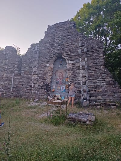 A woman stands in front of a stone structure, possibly ruins, adorned with religious icons and paintings. The structure appears to be made of rough-hewn stones stacked without mortar. A small metal table with some items sits in the foreground, and a stone fire pit is visible to the right. The area is overgrown with grass and weeds. A portion of a tree is visible at the top right. The sky is clear and pale. This location is identified as being in Golovinka, Russia.