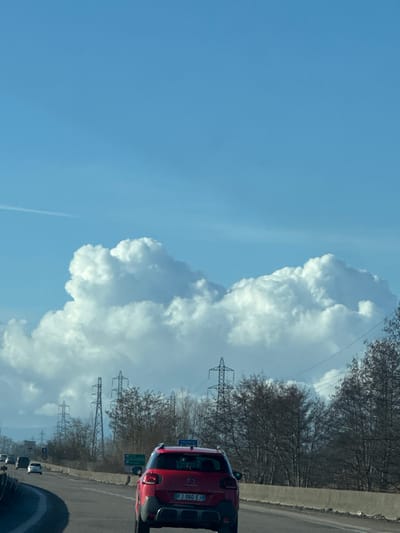 The image depicts a multi-lane highway scene under a clear blue sky with prominent, bright white cumulus clouds. The foreground is dominated by the rear view of a red Citroën C3 Aircross SUV, bearing a French license plate "FJ-060-EJ," traveling in the right lane. Other vehicles are visible further ahead in the left lane.
The highway features a concrete barrier on the right shoulder and a guardrail separating the traffic lanes from the opposing direction. Road signs are present on the right side of the highway; one blue sign clearly indicates "FRANCE," suggesting proximity to the French border. Another green directional sign is partially visible below it, with illegible text.
The landscape adjacent to the highway features numerous deciduous trees, mostly bare, indicating a late autumn or winter season. Extending across the midground and background, multiple large high-voltage electricity pylons are visible, stretching into the distance and silhouetted against the clouds and sky. A faint white contrail is discernible in the upper left portion of the sky. No individuals are visible in the image.
The scene is situated on a highway near Altenheim, Germany, approaching or within the vicinity of the Franco-German border, as indicated by the "FRANCE" sign and the French-registered vehicle.
