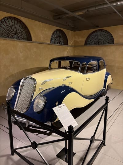 An antique two-toned car, primarily dark blue and cream/beige, is prominently displayed indoors, likely within a museum or exhibition space in Wassenaar, Netherlands. The car's front features a distinctive chrome grille and integrated headlights, with chrome accents visible along its bodywork and door handles. The lower half of the vehicle, including the fenders, is dark blue, contrasting with the cream-colored hood and upper body.
To the right of the car, a black metal barrier or display stand frames the scene. A white rectangular plaque positioned near the car provides descriptive information, with the year "1937" clearly visible, along with the partial text "PANHARD ET LEVASSOR DYNAMIC" and a small logo.
The background consists of light brown/beige walls adorned with multiple circular decorative iron grates, featuring intricate radial and spiral patterns. On the far left, industrial-style silver piping or ducts run vertically along a darker wall section, creating a contrast with the more ornate grates. The car is situated on a light-colored floor, possibly tiled. The overall scene depicts a well-preserved classic automobile showcased under controlled indoor lighting, highlighting its aesthetic and historical value.