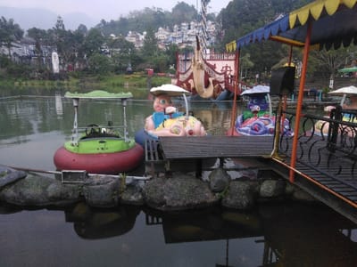 The image depicts an unoccupied section of an amusement park water feature in Ciudad de San José, Costa Rica. In the foreground, dark, still water reflects parts of the scene, bordered by large, moss-covered rocks. A dark wooden dock with black, ornate metal railings extends from the right, partially covered by a blue and yellow striped canopy. Three bumper boats are moored at the dock: a green and red boat with a light canopy, a pink and yellow pig-shaped boat with a white canopy, and a purple and blue octopus-shaped boat with a dark canopy. A wooden ramp leads from the dock into the water next to the character-themed boats. In the midground, a large, elaborate pirate ship ride structure, featuring an ornate bow resembling a sea monster, is visible on the water. The background consists of dense green foliage, an array of multi-colored buildings with fantastical architecture and pointed roofs, and distant mountains under a muted, overcast sky. No individuals are present in the scene.