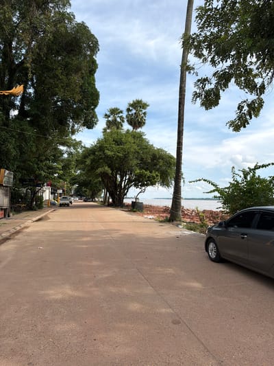The image depicts a street scene in Thakhek, Laos, with a focus on a road lined by trees and adjacent to a river. The road is paved with a reddish-brown material and is mostly empty except for a few parked and moving vehicles. On the left side of the road, there are buildings with awnings and some outdoor seating, suggesting commercial establishments. A large tree with dense foliage dominates the left foreground, partially obscuring a yellow decorative item hanging from its branches.
On the right side of the road, a gray sedan is parked. Beyond the road, a river is visible, with its banks appearing to be undergoing some form of construction or soil disturbance, indicated by piles of reddish earth. Palm trees and other lush greenery are scattered throughout the scene, contributing to a tropical atmosphere. The sky is partly cloudy, with patches of blue sky visible between the clouds. The overall impression is one of a quiet, somewhat unkempt, but picturesque riverside town.