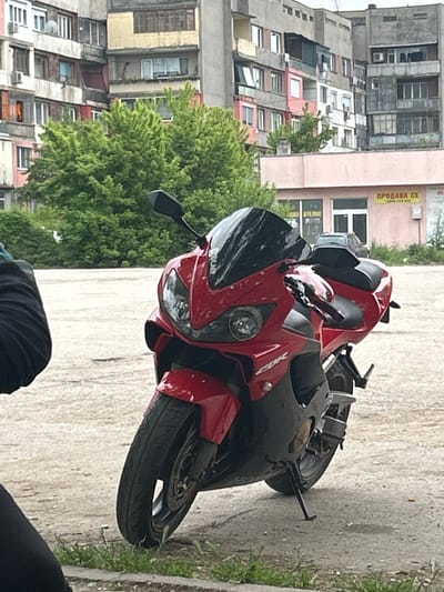 Two motorcycles, one red and one dark, are parked on a concrete surface in front of an urban scene. Buildings with multiple apartments are visible in the background, and trees are scattered throughout. A sign written in Bulgarian, "ПРОДАВА СЕ," is visible on one of the buildings. The image was captured during daylight in Aidemir, Bulgaria. A person wearing black clothing is partially visible in the lower part of the image.