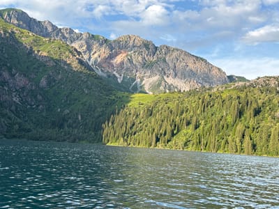 The image shows a wide view of a tranquil lake in the foreground, with its surface rippling under a bright sky. Towering, rugged mountains rise steeply from the far side of the lake. The mountains are covered in dense green forests on their lower slopes, transitioning to rocky, barren peaks higher up. Patches of sunlight highlight the contours of the mountains and the vibrant green of the trees. The sky above is a clear blue, dotted with scattered white clouds. The scene suggests a natural, undeveloped landscape, characteristic of mountainous regions. The location is identified as the city of Arkit, Kyrgyzstan, known for its mountainous terrain and natural beauty.