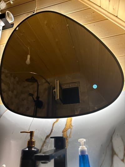 The image displays an interior scene, likely a bathroom, with a prominent, irregularly shaped mirror. The mirror reflects the ceiling, which is constructed of light-colored wooden planks. A single, exposed lightbulb hangs from a white cord in the reflection. Further in the reflection, a shower head and faucet assembly is visible, with a tiled wall behind it. A small window with a white frame is also reflected. On the right side of the mirror's reflection, there is a small, illuminated blue circular symbol.
Below the mirror, on a white countertop with marble-like veining, are two dispenser bottles. The one on the left is black with a gold pump, and the one on the right is clear blue with a white pump. A faucet, appearing to be black, is also present in the foreground.
The overall environment suggests a modern bathroom with wooden accents. There are no people visible in the image. The location within the city of Arkhyz, Russia, cannot be determined from the visual information provided.