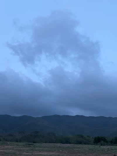 The image shows a wide view of a mountainous landscape under a cloudy sky. The foreground consists of dry, sparse vegetation and soil. Behind this, a band of trees and foliage is visible. The dominant feature is a range of dark, undulating mountains that stretch across the horizon. The sky is a pale blue, with large, diffused clouds that obscure the sun, creating a muted, twilight atmosphere. No people or specific structures are identifiable in the scene. The location is identified as San Antonio, Venezuela.