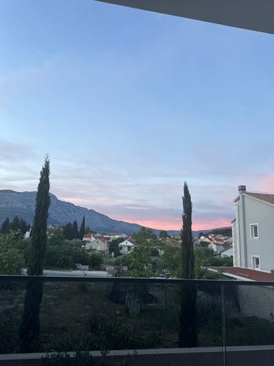 The image depicts a scenic view of the city of Korčula, Croatia, at dusk. In the foreground, a modern balcony railing with a glass panel overlooks a dark, overgrown garden area. Two tall, slender cypress trees stand prominently in the garden. Beyond the garden, a street runs horizontally across the frame, with a few distant objects that might be playground equipment or outdoor furniture.
The middle ground reveals a cluster of residential buildings with red tile roofs, interspersed with lush green trees and foliage. The architecture suggests a Mediterranean style. To the left, a majestic mountain range dominates the background, its slopes appearing rugged and textured.
The sky above is a canvas of soft pastel colors, transitioning from a pale blue at the top to delicate shades of pink and lavender near the horizon, indicative of a sunset or twilight. The overall atmosphere is tranquil and serene, capturing the beauty of a coastal town at the end of the day.