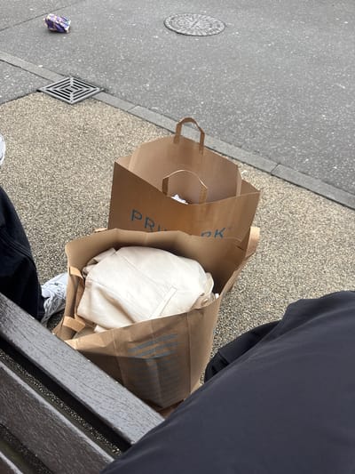 The image shows a close-up, eye-level view of shopping bags placed on a public bench, with a street and sidewalk in the background. Two brown paper shopping bags are the main subjects, one on top of the other. The top bag has the Primark logo visible in blue on its side. Inside the top bag, white or light-colored clothing is visible. The bottom bag also contains clothing, appearing to be folded. The bench is made of dark wood planks. To the left of the bench, a person's legs are visible, wearing dark trousers and white sneakers. To the right, another person's dark clothing is visible. The background consists of a textured gray sidewalk and a darker asphalt street. A metal grate is visible on the sidewalk near the top left of the image, and a circular manhole cover is further in the background on the street. A piece of purple and white litter is also present on the street in the upper left portion of the image. The scene suggests a casual setting, possibly a break or waiting area in a public space within the city of Poole, United Kingdom, given the presence of shopping bags from a well-known retailer.