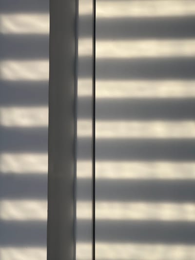 The image shows a close-up of venetian blinds casting striped shadows on a wall. The light is coming from the side, creating alternating bands of light and shadow across the surface. The blinds themselves are not visible, only their effect on the illuminated surface. The colors are muted grays and whites, suggesting a cloudy day or indoor lighting. The scene is abstract and lacks any specific identifying features that would place it in Rotmanka, Poland, or any other location. There are no people, subjects, objects, actions, or interactions depicted. The environment is reduced to a textural interplay of light and shadow on a flat surface.
