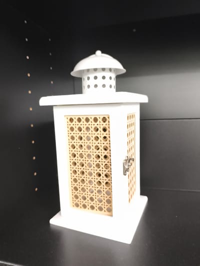 A white lantern with a cane-like patterned window is centered on a black shelf. The lantern has a domed top with ventilation holes. The background is a dark, possibly wooden, shelf with small, lighter colored dots that appear to be imperfections or dust. The lighting suggests an indoor setting, with a shadow cast to the right of the lantern. No people or actions are visible. The image provides no information about the city of Troisdorf, Germany.