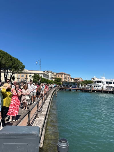 In Desenzano del Garda, Italy, on a sunny day, a long queue of people waits along a waterfront promenade. The people, dressed in casual summer attire, are lined up on a paved walkway with a metal railing. The walkway overlooks a body of turquoise water, likely Lake Garda, where a white ferry boat is docked at a pier. In the background, several buildings with traditional Italian architecture are visible, including a multi-story building with cream-colored facades and arched windows. Trees line the promenade, and the sky is a clear, bright blue. The scene suggests a popular tourist location, possibly awaiting transportation or an attraction.