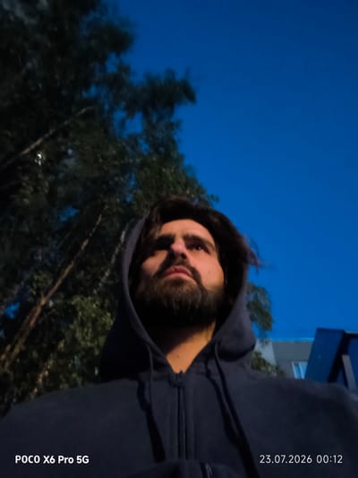 A low-angle, medium shot captures a man wearing a dark blue hooded sweatshirt, looking upwards. His face, framed by dark hair and a beard, is illuminated by an unseen light source. To the left, the dark foliage of trees with visible branches is silhouetted against a deep blue twilight sky. In the background, on the right, a portion of a blue structure is visible. The lower portion of the image displays a watermark in the bottom left corner reading "POCO X6 Pro 5G" and a date and time stamp in the bottom right corner reading "23.07.2026 00:12". The scene is consistent with an outdoor setting in Kogalym, Russia, during twilight.