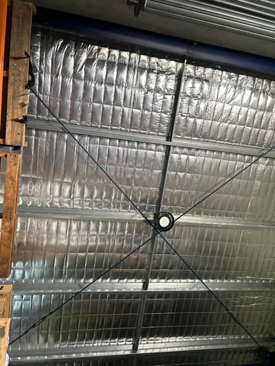The image displays the interior ceiling of an industrial or storage facility located in Stanwell Tops, Australia, viewed from below looking upwards. The ceiling consists of a metallic corrugated roof structure supported by horizontal galvanized steel purlins. Below the roof, a silver, quilted thermal insulation material is installed between the purlins. A single circular LED high-bay light fixture, black with fins, is centrally suspended from the ceiling structure. Four blue tension cables or bracing wires extend diagonally from the perimeter towards the center, converging near the light fixture. On the far left, a vertical wooden structural beam is partially visible, with a dark, shredded material attached to its upper section. Above the insulation in the upper portion of the frame, a dark blue horizontal beam is present, beneath what appears to be a section of a metallic roller door mechanism. No individuals, explicit actions, or interactions are visible in the scene.