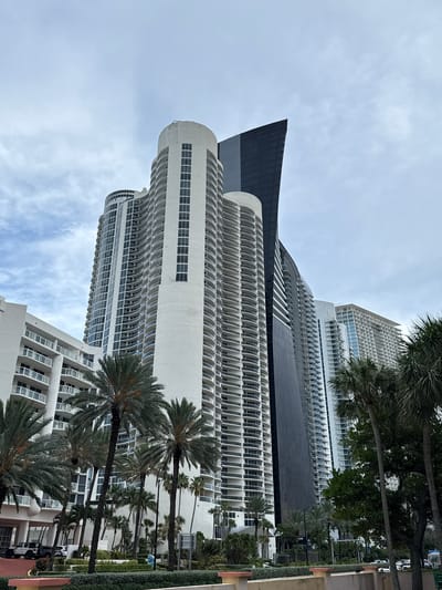 The image shows a cluster of modern, high-rise buildings, likely residential condominiums, against an overcast sky in Sunny Isles Beach, Florida, USA. The buildings are characterized by their curvilinear white facades with numerous balconies and dark-tinted windows. One particularly striking building on the right features a sharp, angled, dark-colored facade, contrasting with the softer lines of the surrounding structures. Palm trees are interspersed throughout the foreground and middle ground, some with their fronds obscuring parts of the buildings. In the lower left corner, the corner of a white, multi-story building with balconies is visible. Below the buildings, there are signs of urban infrastructure, including paved areas, landscaping with manicured hedges, and what appear to be parked vehicles, including two SUVs. The overall impression is of a contemporary, upscale coastal urban environment.