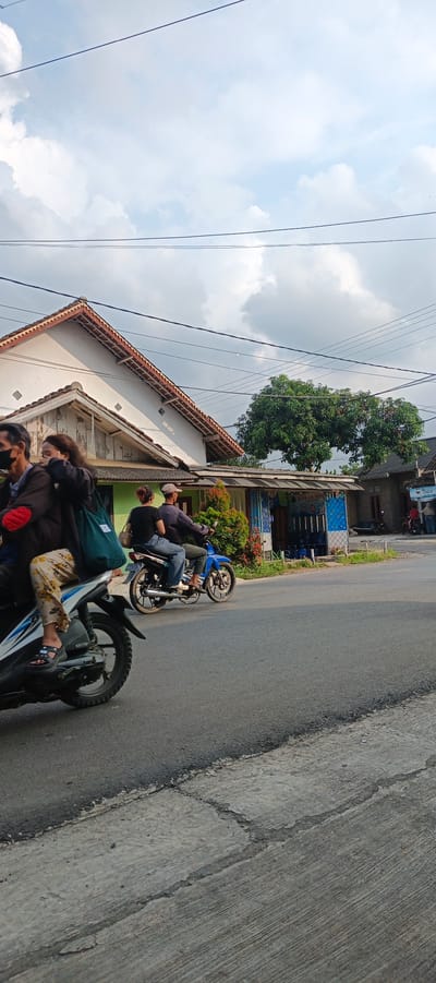 In Sukarame, Indonesia, on a paved road, two motorcycles are in motion. The foremost motorcycle, partially visible on the left, carries two individuals, one driving and another seated behind. The driver is masked and wears a dark jacket and patterned pants. The passenger is also masked, wearing a dark jacket and carrying a teal bag. They are traveling from left to right across the frame.
Behind them, a second blue motorcycle is also moving from left to right, carrying two people. The driver, wearing a dark long-sleeved shirt and a light-colored cap, is operating the vehicle. The passenger, in a black t-shirt and blue jeans, is seated behind.
The background features a building with a white facade and a tiled roof, partially obscured by a large green tree. Further to the right, another building with a green exterior and a blue-patterned facade is visible, with a sign that reads "DEPO AIR MINUM". The sky above is partly cloudy with a bright, hazy sun. The road surface is asphalt, with concrete on the side.