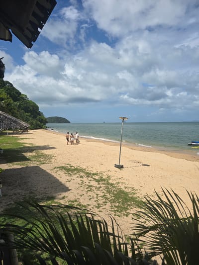 The image shows a beach scene in Ban Khlong Hia, Thailand. The sandy beach is visible, with ocean water along the shoreline. There are a few people standing on the beach near the water. A tall, gray light pole is standing upright on the beach. Palm tree leaves are in the foreground. Green trees can be seen behind the beach. The sky is visible with clouds. Part of a building is in the lower-left corner.