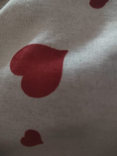 The image displays a close-up, abstract view of a white fabric with a repeating pattern of red hearts. The fabric appears to be textured, with visible weave lines. The hearts are of varying sizes and orientations, with some appearing to be partially obscured by folds in the material. One large heart is prominently featured in the center-left of the image, while a smaller heart is visible in the lower-left corner. Another heart is partially visible in the upper-right corner. The lighting creates shadows and highlights on the fabric, emphasizing its texture and the curves of the hearts. There is no information available about the location in Dorkovo, Bulgaria, as the image solely focuses on the fabric pattern.