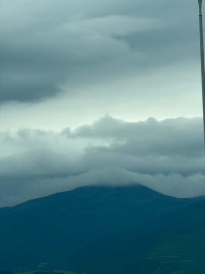 The image shows a view of mountains and clouds in Tedotsminda, Georgia. The mountains are dark green and the clouds are white and gray. The sky is overcast. A pole crosses the top of the frame.