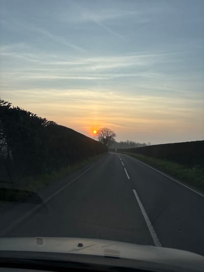 The scene depicts a rural road, observed from within a vehicle looking forward through its windshield, located in the vicinity of Baschurch, United Kingdom, during sunrise. The sky displays light blue at the top, transitioning to an orange and yellow gradient at the horizon where the sun is visible, partially diffused by the leafless branches of a prominent tree. The asphalt road extends into the distance, featuring a continuous white line on its left edge and a broken white centerline, and curves slightly to the right. To the left of the road, a tall, dark green hedge runs parallel, behind which the silhouetted bare tree stands. On the right, a lower, dark hedge abuts the road, with an expanse of dark, possibly cultivated, land beyond it, and a distant tree line merging with atmospheric haze. No persons or discernible activities are depicted.