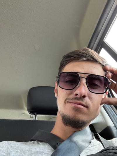 The image is a close-up, selfie-style photograph taken inside a vehicle. The subject is a young man wearing sunglasses with a dark, gradient tint and a black metal frame. He has brown hair styled casually and a short beard and mustache. He is wearing a light grey t-shirt and a grey seatbelt is visible across his chest. His right hand is resting on his head, near his temple, as if he is leaning his head or adjusting his hair. The background of the image shows the interior of a car, including a headrest of a dark grey or black seat and the ceiling of the car, which appears to be a light beige or grey textured material. Natural light is entering from a window on the right side of the frame. The specific location in Biaga, Bulgaria is not discernible from the image.