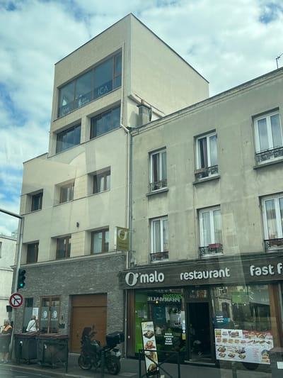 This is a street-level, eye-level, medium-range, eye-level shot of a street in Alfortville, France, on a partly cloudy day. The scene features a modern building with a grey brick lower level and a beige upper section with large windows. To the right, an older, grey building with multiple windows is visible. A sign for "O'malo restaurant fast food" is prominently displayed above the entrance of a restaurant, which features various promotional posters for food items. A green traffic light is visible on the left, along with a traffic sign indicating a no-entry zone and two large trash bins. A man in a reflective vest stands near the bins, and a black motorcycle is parked in front of a wooden garage door on the lower level of the modern building. The windows of the modern building display company names such as "ICA", "SKY INGENIERIE", and "BMI".
