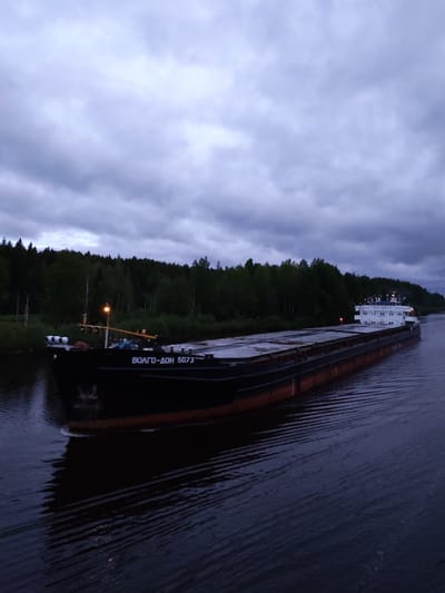 A large river barge, identified as "ВОЛГО-ДОН 5073" in Cyrillic script, is moving through a waterway. The vessel is dark in color with a red-brown waterline stripe. It is situated on dark, rippled water, likely a river or canal. The background features a treeline, suggesting a rural or natural environment. The sky is overcast with heavy, dark clouds, indicating either dusk or a storm approaching. The photo was taken at Александровское, Russia.
