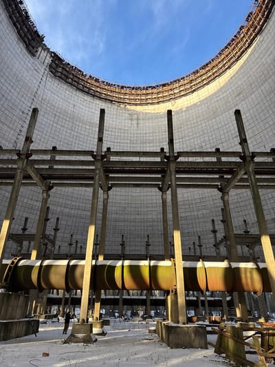 An upward-angled view captures the interior of a large, decaying structure. A blue sky is visible through a massive circular opening. Metal scaffolding and concrete structures dominate the scene, suggesting industrial or infrastructural origins. A central, cylindrical object with visible banding is highlighted by sunlight, casting shadows on the surrounding surfaces. The photo's perspective creates a sense of depth and scale.
