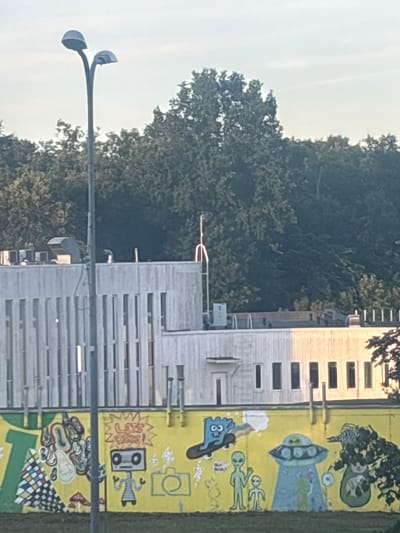 The image shows a section of a building with a yellow mural on its side. The mural depicts various cartoonish figures and objects, including aliens, a UFO, a robot, and what appears to be a stylized mouth. There are also abstract shapes and patterns. The building itself has a modern architectural style with horizontal window slats. In the foreground, a street lamp with two curved arms and lights is visible. Trees and a bright sky suggest an outdoor setting, possibly during the daytime. The mural’s colorful and whimsical nature might evoke a sense of fun or playfulness. Text is visible on the mural, including "LET'S DRIVE!" above the robot image, and what appear to be numbers or symbols below it. The scene is vertical, suggesting the photo was taken with the camera rotated. The location is Jõhvi, Estonia.