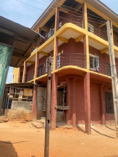 The scene depicts an exterior view of a three-story building with reddish-brown walls and prominent yellow-painted beams and balcony railings, situated in Iyiowa Odekpa, Nigeria. The building features multiple windows, some open, and balconies with dark metal railings on each floor. A ground-level entrance displays a partially visible sign with text "MENT" or "CEMENT" and phone numbers "0704307064", beneath which stacked white sacks are visible, indicating a commercial establishment. To the left, a single-story structure with a corrugated metal roof and partially green-painted walls is visible, including a "Coca-Cola" sign. The ground is unpaved, consisting of dry, reddish-brown earth. A weathered metal pole with circular holes stands vertically in the mid-ground. The sky is clear blue. No individuals are present, and no actions or interactions are depicted.