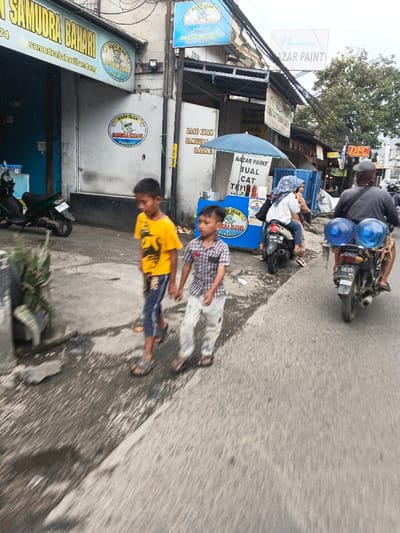 The image captures an active street scene in Dayeuhkolot, Indonesia. In the foreground, two young boys walk left-to-right on an uneven concrete path. The boy on the left wears a yellow t-shirt and dark shorts, while the boy on the right, in a plaid shirt and light pants, holds his hand. Adjacent to them, on the asphalt road, a dark motorcycle travels right-to-left. The driver, wearing a dark shirt and head covering, transports two large blue water dispenser bottles secured to the rear, with a license plate displaying "D 6639". A female passenger in a white shirt and blue hijab is seated behind the driver.
On the left side of the scene, behind the walking boys, a row of commercial buildings features various signage. A prominent storefront displays "SAMUDRA BAHARI" and "BASO IKAN" (fishball) logos. Further right, a blue vendor stall with a blue umbrella is positioned at the roadside, marked with "NAZAR PAINT JUAL CAT" (paint for sale) and "BASO IKAN SAMUDRA BAHARI" text. A parked green motorcycle is partially visible on the far left. Overhead, numerous utility lines crisscross against an overcast sky. The ground on the left side of the path appears damp or wet in sections.