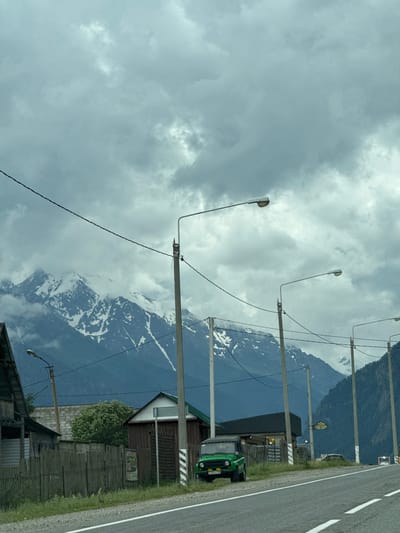 In Верхняя Теберда, Russia, a cloudy day looms over a landscape featuring snow-capped mountains. The foreground shows a road running alongside a wooden fence, with a few scattered buildings and a green SUV parked on the side. Overhead, power lines crisscross the sky, supported by concrete poles, with streetlights mounted on some of them. The mountains, partially obscured by clouds, exhibit rugged terrain with patches of snow. The overall scene depicts a rural or semi-rural setting in a mountainous region.