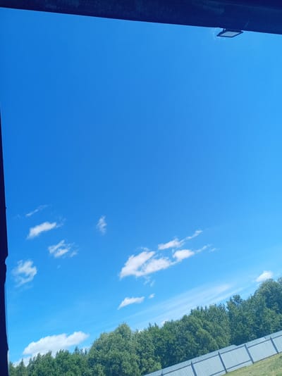 The media file shows a bright, clear blue sky with scattered white clouds. In the foreground, a dark structure, possibly a roof overhang or window frame, partially obscures the top of the image. To the right of the frame, a modern light fixture is attached to this structure.
Below the sky, a dense line of green trees forms the horizon. In the lower right corner, a grey metal fence runs diagonally across the scene. The grass in front of the fence appears green and slightly sloped.
The overall impression is of a sunny day, likely late morning or early afternoon, given the brightness and the clear sky. The setting suggests an outdoor environment, possibly rural or suburban, in Belarus, as indicated by the location context. There are no people visible in the image. The atmosphere is calm and serene, with the natural elements of sky, trees, and a fence dominating the frame. No text is visible in the media file.