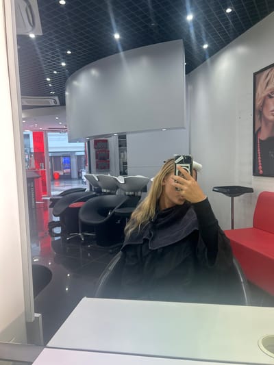 The image shows the interior of a hair salon in Leers, France. A woman sits in a salon chair, taking a selfie with her phone. She has blonde hair and is wearing a black garment with a dark towel draped over her shoulders. Her hair appears to be undergoing a treatment of some sort. The salon features a modern design with black and white walls and ceiling, accented by a diamond grid pattern. Red seating is visible. Through the window, elements of the city streetscape are visible, including street signs. "LEERS COMMANDEZ ICI" is visible in reverse on the window.