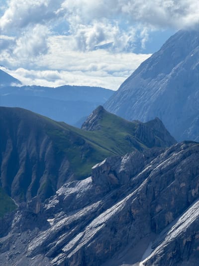 The image captures a mountainous landscape with layers of peaks and valleys under a cloudy sky. In the foreground, rugged, gray rock faces with sharp, angular formations are visible. Further back, rolling green hills with patches of grass transition into rocky outcrops. The background features progressively lighter blue mountain ranges, fading into the hazy sky. The clouds are white and scattered, with some appearing dense and others wispy, allowing glimpses of a bright sky. The overall impression is of a vast, natural, and somewhat dramatic alpine environment. There are no people or man-made structures clearly identifiable in the image, focusing solely on the natural topography.