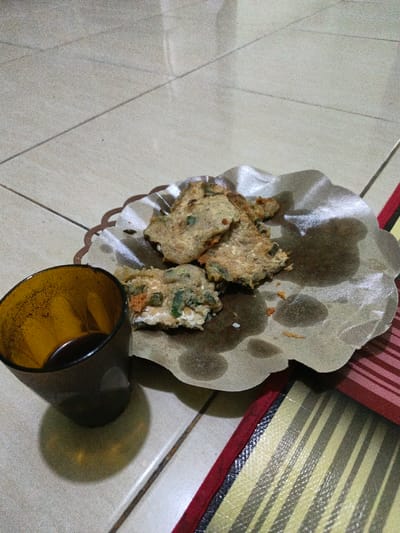 The scene depicts food and a beverage set on a tiled floor in Lagadar, Indonesia. In the foreground, a dark amber-colored glass containing a dark liquid is positioned on the lower left. Adjacent to it, in the center, several pieces of fried food, consistent with fritters, rest on a crinkled, brown paper liner. The paper exhibits multiple dark, circular oil stains. The food items are light brown and irregular in shape, displaying visible green and orange inclusions within their texture. One piece appears fractured, exposing its internal structure. A few scattered white particulates are present on the paper. The paper liner is situated within a partially visible brown, scalloped-edge holder. The immediate surface is a light-toned tiled floor with visible grout lines. A section of a striped textile, featuring alternating light and dark horizontal bands with a red-brown edge, is partially visible in the lower right. No individuals or actions are depicted.