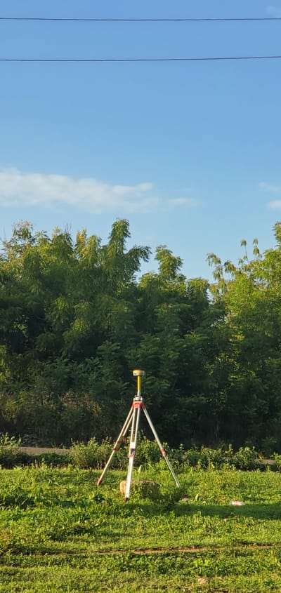 The image captures an outdoor scene, likely a rural or semi-rural area in Iganna, Nigeria, with surveying equipment set up in a grassy field.
In the foreground and midground, a red and white striped tripod stands firmly on the vibrant green grass, supporting a yellow-capped surveying device, which appears to be a GNSS receiver or similar land measurement instrument. The legs of the tripod are extended, indicating it is in active use for surveying. The ground is covered in dense, overgrown green grass and various plants. A white and red plastic bottle is visible lying on the grass behind the surveying equipment.
The background features a thick line of lush, dark green trees and bushes, suggesting a forested area or dense vegetation bordering the field. Above the tree line, a clear, bright blue sky dominates the upper portion of the image, with a few wispy white clouds scattered across it. Two distinct dark power lines stretch horizontally across the top left of the sky, indicating the presence of utility infrastructure nearby.
The scene suggests a sunny day, likely in the morning or late afternoon, given the bright illumination. No people are visible in the image, implying the equipment might be set up for a data collection session or is temporarily unattended. There is no discernible text visible on any of the objects or in the scene.