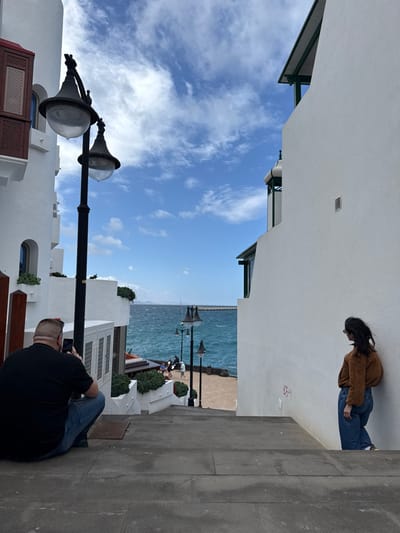 An outdoor scene in Playa Blanca, Spain, depicts a descent via concrete steps towards a turquoise waterfront under a blue sky with scattered white clouds. White-washed buildings flank the steps; the building on the left features a dark brown wooden balcony, while the one on the right has a smooth facade with a small vent.
In the immediate foreground, a male individual is crouched on the steps, facing away from the viewer. He wears a black t-shirt and blue jeans, with sunglasses perched on his head, and is holding a smartphone to capture an image of the scene ahead. To his right, a female individual with dark hair, wearing a brown long-sleeved top and blue jeans, leans against the white wall of the building, looking towards the left side of the frame.
Further down the steps, a black lamppost with two ornate light fixtures stands prominently. In the midground, at the base of the steps, a paved promenade borders the sea. Several indistinct figures are visible on the promenade, some appearing to be seated. The sea is moderately choppy, and a distant breakwater or pier extends from the right shore. Beyond the sea on the horizon, landforms are visible under the same sky. Additional smaller lampposts are visible along the promenade. Green foliage in planters and on balconies contributes to the scene.