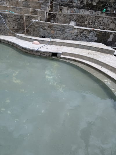 The image shows a terraced pool with stone walls and concrete steps in Panamik, India. The pool is filled with murky, greenish-blue water. On one of the concrete steps, there is an orange, dome-shaped object and a blue hose. The stone walls are constructed of flat, irregular stones stacked in horizontal layers. The environment appears to be outdoors, with natural light illuminating the scene. The image does not contain any people or obvious signs of recent activity. The architecture suggests a man-made structure, possibly a public bath or a natural hot spring facility, given the location in Panamik which is known for its hot springs.