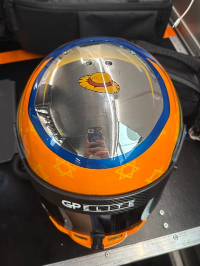 The image shows a close-up of a racing helmet with a reflective visor. The helmet is painted orange and blue and features a stylized depiction of a straw hat on the top. The helmet is decorated with multiple Star of David symbols. The helmet is positioned on top of a black metal box with a silver-colored surface, and the reflective visor shows a blurry reflection of a person holding a black cell phone. This image was taken in Montmeló, Spain, at coordinates 41.57 latitude and 2.259 longitude. The scene is likely set at the Circuit de Barcelona-Catalunya race track, as the helmet features "GP Elite" written on it.