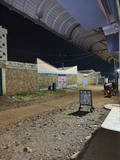An outdoor shot at night shows a dirt road lined with brick buildings. Each building features a pitched roof and a brightly painted door. A sign for "Cyber Services, Movie Series" stands along the road. Further down, two people and a moped are visible. Overhead, a corrugated metal roof provides a partial covering over the foreground. Streetlights glow in the distance against the dark sky. A tall building stands on the left.