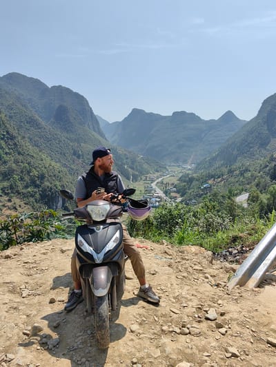 A man sits on a moped on a rocky, dirt-covered incline overlooking a valley, likely in the Phố Bảng Commune, Vietnam, based on the mountainous terrain and the valley community visible in the background.
The man, fair-skinned with a beard and wearing a navy baseball cap, gray t-shirt, and a dark vest, is seated on the moped, looking off to his left. He holds a cell phone in one hand. His legs are khaki pants, and he wears gray sneakers. A purple helmet is seen resting near the moped's handlebars.
The moped is a dark color, mostly black and gray, and is positioned on the dirt path. The foreground is rocky and sparsely vegetated with some small plants. A metal guardrail is visible at the edge of the path on the right side of the image.
The valley beyond features a town or community with buildings and a road winding through it. Mountains covered in green vegetation rise in the background under a clear, blue sky. The overall scene suggests a scenic overlook on a hillside.