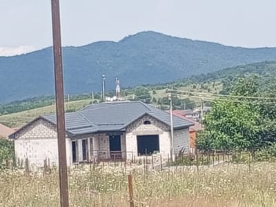 An outdoor, daytime, medium shot captures a partially constructed house with a gray, tiled roof, situated in a rural area. The house is made of white cinder blocks, with visible openings for windows and doors. Two figures stand on the roof, one appearing to be holding something aloft. A tall, brown wooden post dominates the left foreground, obscuring part of the scene. In the background, rolling hills covered in dense green vegetation and trees lead up to a large, dark blue mountain range under a pale blue sky. A field of wildflowers and dry grass occupies the area in front of the house. The overall setting suggests a construction site in a mountainous, undeveloped region. This location is identified as Элистанжи, Russia.